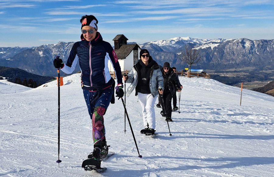 Gruppe bei einer Schneeschuhwanderung vor Bergkapelle und verschneitem Alpenpanorama